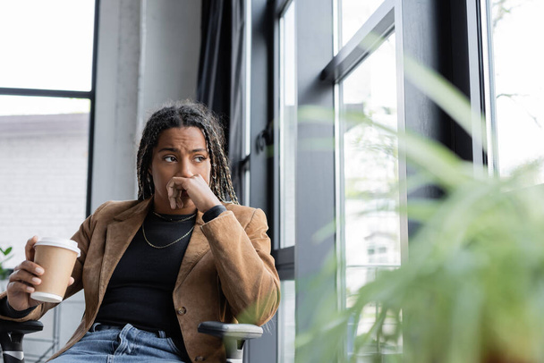 Ανήσυχη Αφροαμερικανή επιχειρηματίας με σακάκι κρατώντας πακέτο ποτό κοντά στο παράθυρο στο γραφείο  - Φωτογραφία, εικόνα