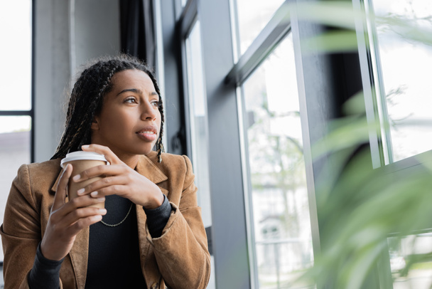Femme d'affaires afro-américaine tenant un café pour aller près de la fenêtre dans le bureau  - Photo, image