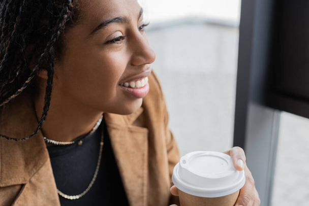 Smiling african american businesswoman in jacket holding paper cup in office  - Fotó, kép