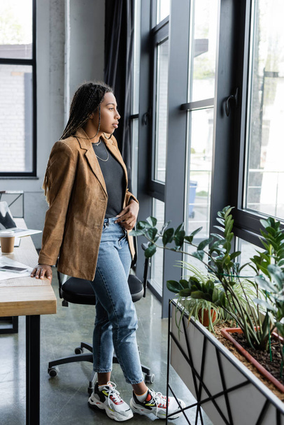 Side view of african american businesswoman in jacket and jeans looking at window in office  - Φωτογραφία, εικόνα