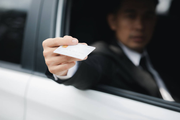 Un Asiatique en costume classique reste dans la voiture, ouvre la fenêtre, remet sa carte de crédit pour payer l'essence, voyage en voiture, conduite sûre, respect des règles de circulation. - Photo, image