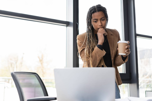 Focused african american businesswoman holding coffee to go and looking at laptop in office  - Foto, imagen
