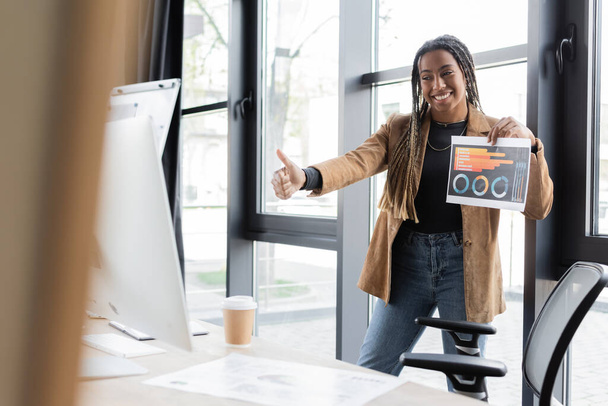 Африканская американская бизнесвумен показывает как жест и держит бумагу во время видеозвонка на компьютере в офисе  - Фото, изображение