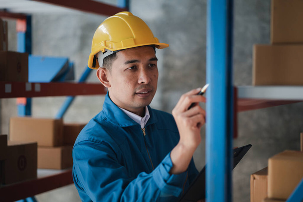 Asian man wearing protective clothing working in a warehouse, he is checking stock, wearing protective clothing while working at a large warehouse according to company policy and worker safety. - 写真・画像