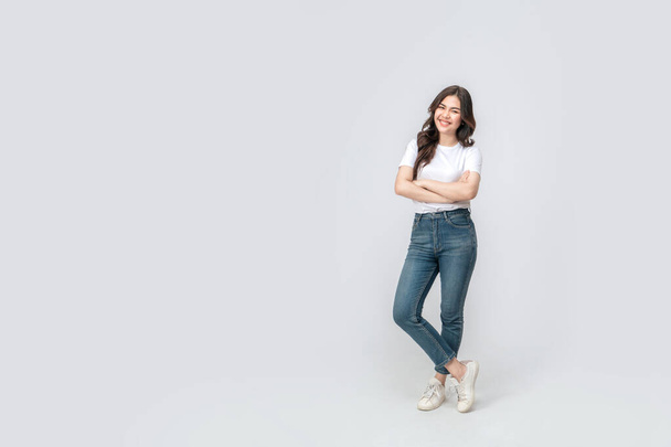 Pleine longueur de femme asiatique confiante souriant dans une tenue décontractée dans un t-shirt blanc et un jean, debout avec son bras et sa jambe croisés, avec un sourire sur son visage sur fond blanc isolé. - Photo, image