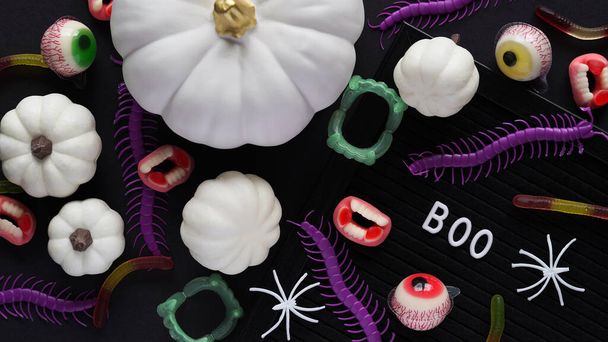 Pumpkins, cauldrons, pots, worms, jaws, spiders, eye-shaped candies and a text board with the inscription BOO on a black background, top view. A collection of items for a Halloween party - Foto, Bild