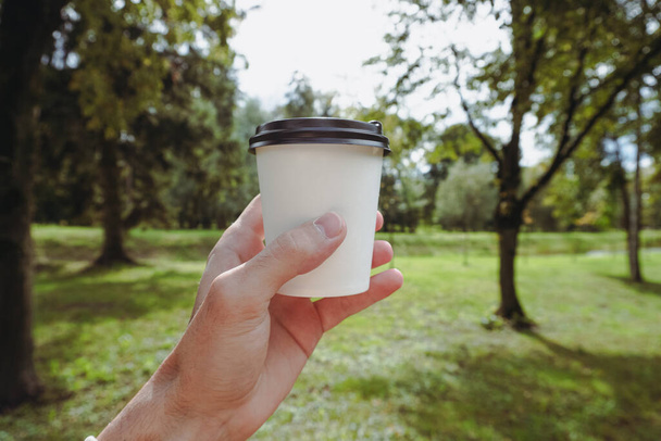 Hand holding paper coffee cup. Paper white cup for coffee on a background of grass. A man holds a mockup of a paper cup for coffee and tea outdoors. Close-up view of paper coffee cup in hand. - Фото, изображение