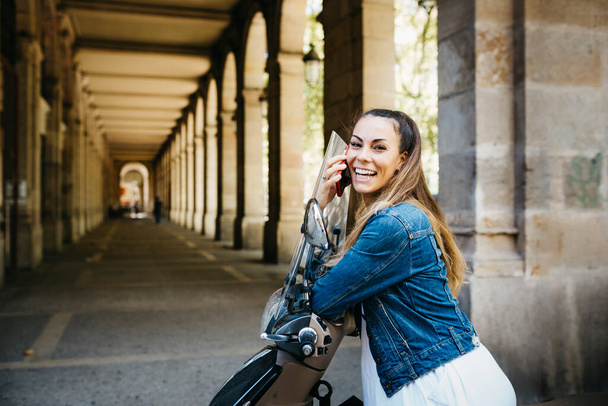 Mladá usměvavá žena na motorce mluví na červeném smartphonu - Fotografie, Obrázek