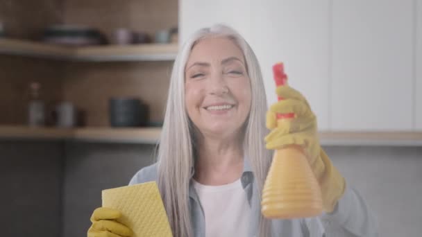 Hravá žena hospodyňka starší běloška babička uklízečka nosí žluté gumové rukavice pózující s vlhkou houbou a postřikovač stříkající prací prášek ve vzduchu se baví přípravou na čištění kuchyňské práce - Záběry, video