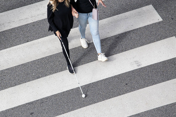 Σκηνή μιας τυφλής γυναίκας που περπατούσε στο πέρασμα της ζέβρας και την βοηθούσε ένα άλλο άτομο χρησιμοποιώντας το λευκό μπαστούνι της. Βοήθεια στα πρώτα στάδια της τύφλωσης - Φωτογραφία, εικόνα