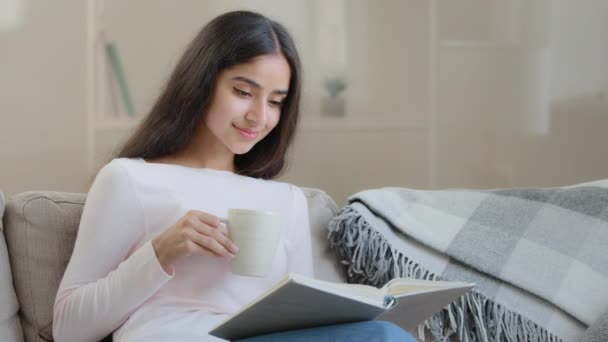 Arabska brunetka kobieta uczeń student siedzieć na kanapie czytać papier książka w twardej oprawie romantyczna literatura opowieść uczyć się nowego języka przygotowując do egzaminów domowych trzymać filiżankę kawy kulturalny wypoczynek w domu - Materiał filmowy, wideo