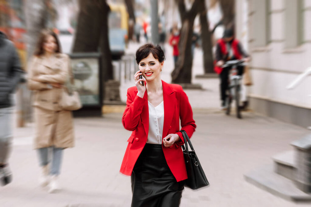 Νεαρή με αυτοπεποίθηση επιχειρηματίας σε επίσημο ένδυμα, κόκκινο σακάκι μιλάει στο smartphone, ενώ το περπάτημα στο δρόμο της πόλης. Τηλεπικοινωνίες και κινητό δίκτυο για επιχειρηματική ιδέα. Γυναικείο επιχειρηματικό στυλ. - Φωτογραφία, εικόνα