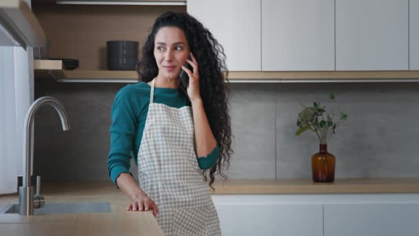 Csinos arab göndör hajú nő anya háziasszony visel kötény állvány konyhai beszélgetés mobiltelefon barátságos beszélgetés hívás szállítási élelmiszer szolgáltatás rend otthonról töltsön szabadidőt egyedül - Felvétel, videó