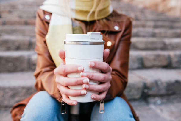 Felismerhetetlen fiatal nő termo csésze forró elviteles kávéval a város utcáin, a kőlépcsőn ülve. - Fotó, kép