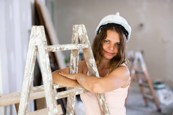 Young attractive girl in beige dress and protective helmet poses on stepladder in a room being renovated - Zdjęcie, obraz