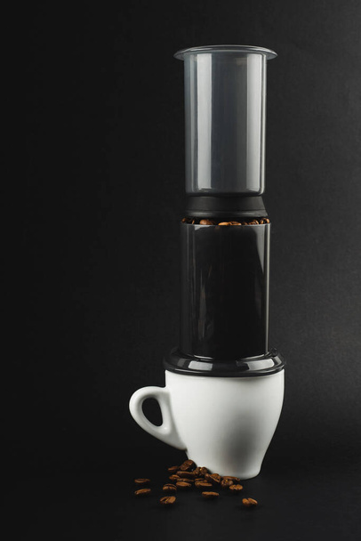 Aeropress with coffee beans, white espresso cup on black background. Aeropress is a device for brewing coffee - Foto, Imagem