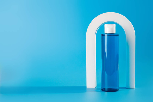 Micellar μπουκάλι νερό ή τονωτικό για περιποίηση προσώπου. Καλλυντικό προϊόν περιποίησης σε γυάλινη απόχρωση στο μπλε φόντο των λευκών τόξων. Αντιγραφή χώρου - Φωτογραφία, εικόνα