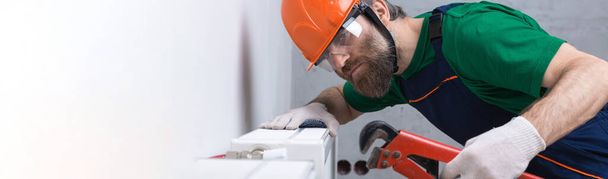 Um encanador masculino instala um radiador no sistema de aquecimento de um apartamento. Um tipo de macacão e uma chave de gás.. - Foto, Imagem