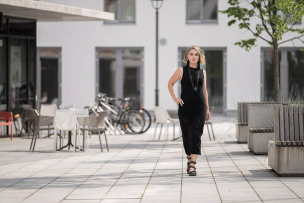Vilnius Old Town Street in Lithuania. Beautiful White Caucasian Adult Woman is Wearing Black Dress and Bijouterie on Neck. Shops and Restaurant in Background with Chairs Outside - 写真・画像