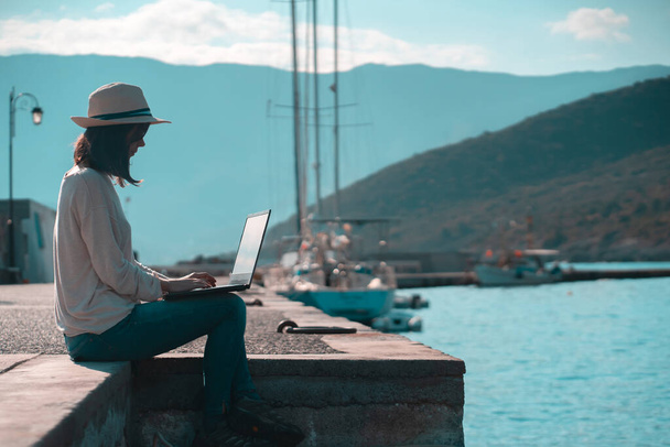 Uma jovem de chapéu está sentada no cais e trabalhando, digitando em um teclado de laptop em um dia ensolarado contra um belo fundo de uma paisagem marinha com iates ancorados na baía. Mulher trabalha e viaja - Foto, Imagem
