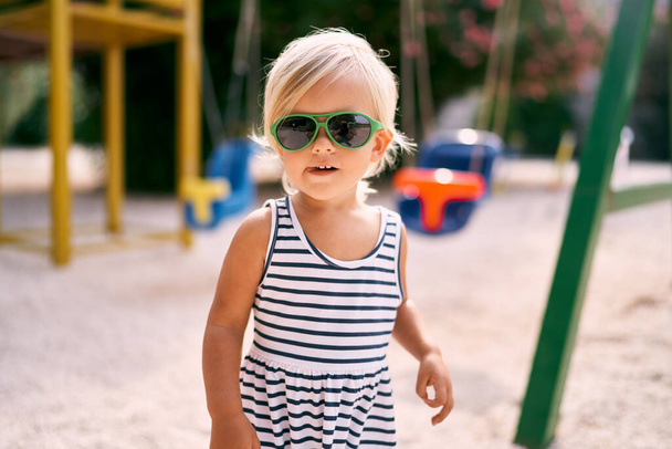 Маленькая загорелая девочка в солнечных очках на детской площадке. Портрет. Высокое качество фото - Фото, изображение