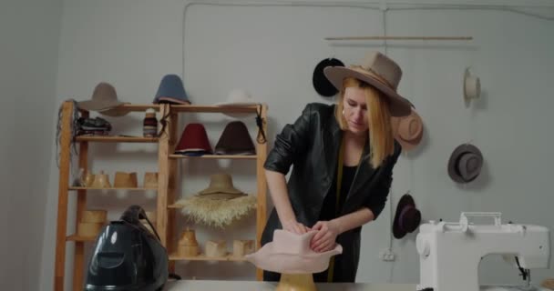 Woman makes shape of female hat with hands using wooden mold at table. Experienced milliner works with headwears in workshop closeup - Кадры, видео
