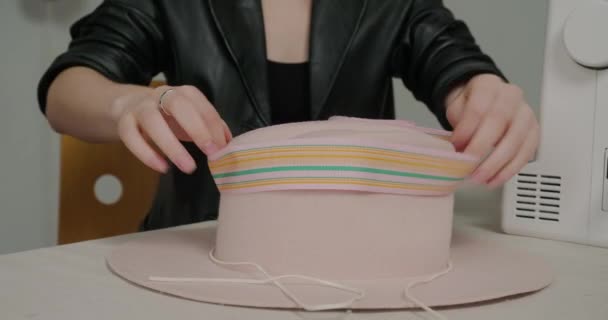 Milliner laittaa leveä värikäs vanteen huopahattu istuu pöydässä lähellä ompelukone. Nainen mustassa nahkatakissa koristaa päähineet lähikuva - Materiaali, video