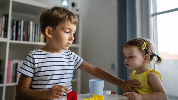 One preschool boy playing at home with science experiments while his little toddler sister interrupts him and he trying to prevent her to take his science equipment childhood siblings growing up concept copy space - Foto, Bild