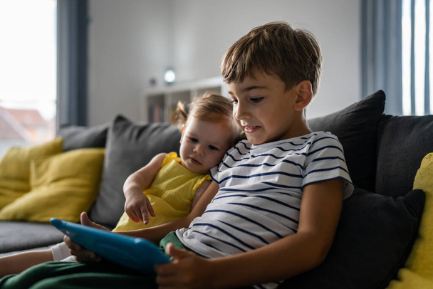 Um menino pré-escolar sentado com sua irmãzinha no sofá em casa olhando para tablet digital jogando jogos aprendendo irmãos crescendo conceito - Foto, Imagem