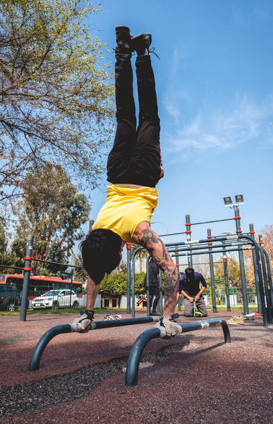 Zdrowy i sprawny młody Latynos z wytatuowanym rękawem, żółtą koszulą i rękawiczkami treningowymi wykonujący ćwiczenia gimnastyczne (odwrócona pozycja) na ulicznym parku treningowym w słoneczny dzień - Zdjęcie, obraz