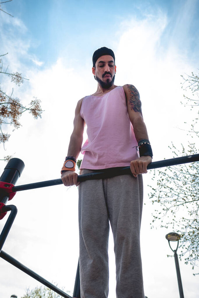 Fit and handsome athletic latin guy with cap and colorful tattoo doing a calisthenic exercise routine (performs a muscle-up) in an street workout park in a sunny day - Photo, Image
