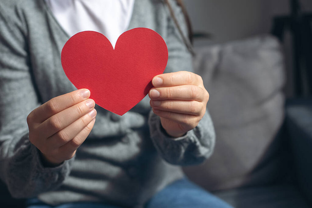 World heart day, world health day, CSR responsibility, adoption foster care home, all lives matter, no to racism concept. Close-up of young female holding small red paper heart sitting on comfy couch - Foto, Imagem