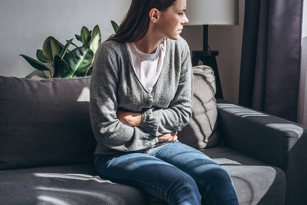 Δυσάρεστη νεαρή γυναίκα 20 ετών κάθεται σε καναπέ στο σαλόνι στο σπίτι, πάσχει από πόνο κοιλιακών κράμπες, αναπνέει βαριά, κρατώντας το στομάχι, γαστρίτιδα, κοιλιακή, κορύφωση έννοια - Φωτογραφία, εικόνα