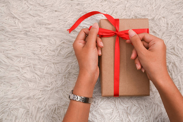Egy nő keze piros szalagot köt egy kézműves karácsonyi ajándékdobozra fehér szőrszőnyegen. A karácsonyi ünnep koncepciójának előkészítése. - Fotó, kép