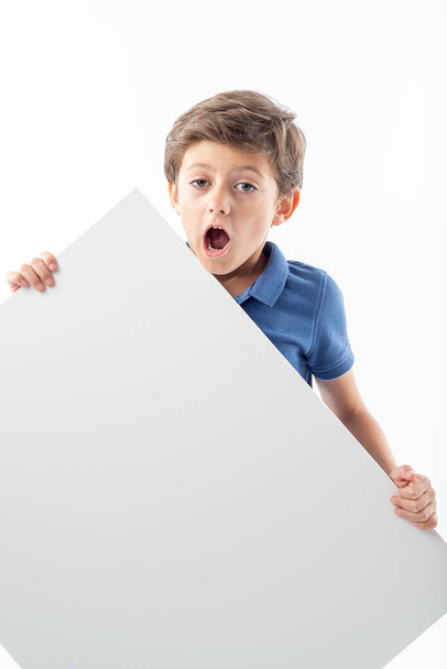 Caucasian boy holding white sign with space for text and looking at camera with surprised expression, white background. - Fotoğraf, Görsel