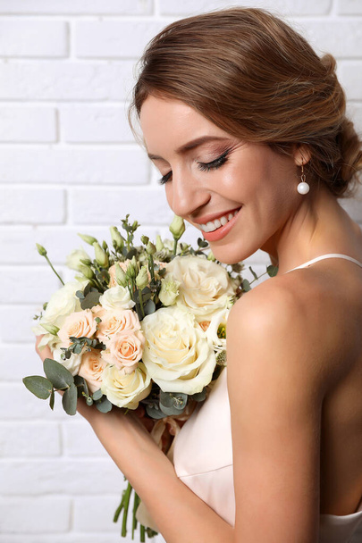 Young bride with elegant hairstyle holding wedding bouquet near white brick wall - 写真・画像