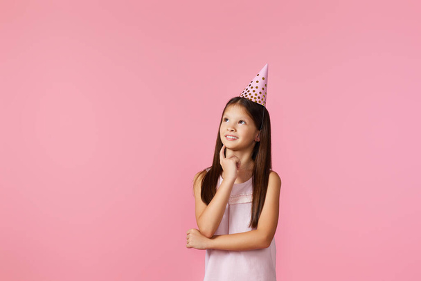 little girl with party cone making wish on her birthday on pink background. copy space - Zdjęcie, obraz
