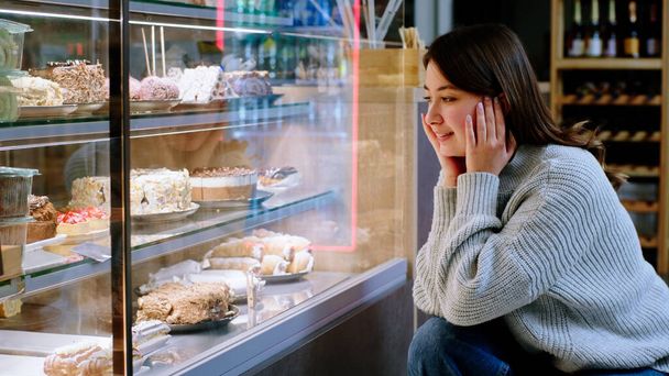 At the cozy coffee shop happy lady very hungry looking at the showcase fridge to choose her favourite dessert. - Foto, imagen