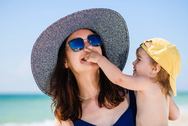 Güneş şapkalı ve gözlüklü anne ve kız plaj şezlongunda uzanıp oynuyorlar. Deniz arka planında dinleniyorlar. Mutlu aile tatilleri. - Fotoğraf, Görsel