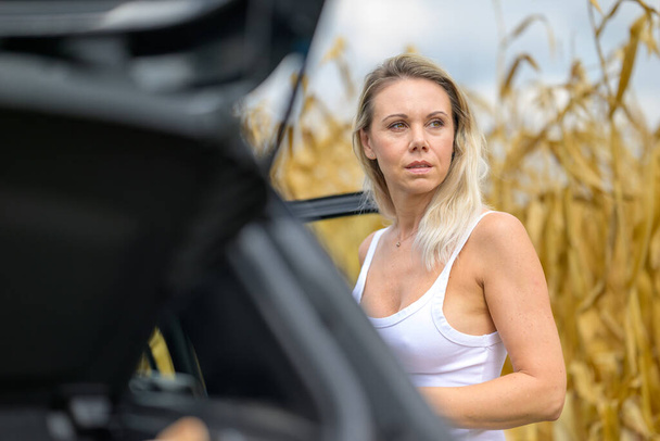 Vrouw staat bij een auto met een doordachte blik vanwege een autopech voor een maïsveld - Foto, afbeelding
