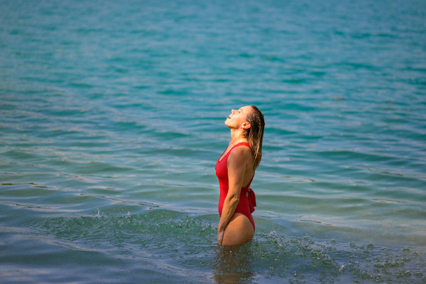 Hermosa mujer rubia atractiva con el pelo mojado en un traje de baño rojo está de pie en el agua con los ojos cerrados y disfrutando del agua - Foto, imagen