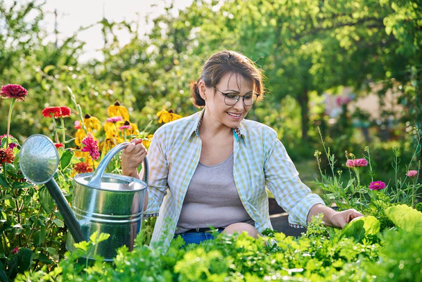 Outdoor portrait woman with watering can in vegetable garden in summer season. Smiling middle aged female looking at camera, beds with vegetables herbs flowers background - Foto, Imagen