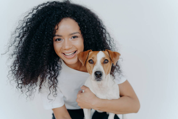 Close up shot of beautiful happy Afro woman with bushy curly hair, embraces favourite dog and have fun together at home, expresses love to jack Russell terrier puppy, isolated over white background
 - Фото, изображение