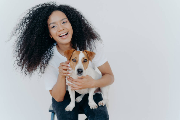 Portret radosnej kręconej dziewczyny pieszczącej swojego psa, radującej się kupując Jacka Russella Terriera, uśmiecha się szeroko, bawi się ze zwierzętami, nosi luźne ubrania, odizolowane na białym tle, cieszy się dobrym dniem - Zdjęcie, obraz