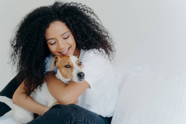 Happy African American woman expresses love to dog, embraces pet, dressed in casual wear, sit on comfortable bed in bedroom, sleeps with domestic animal. Jack russell terrier with owner in morning - Φωτογραφία, εικόνα