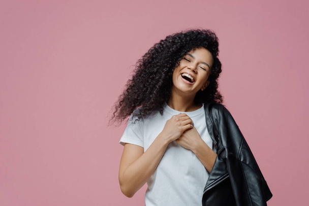 Overjoyed dark skinned woman keeps hands together and laughs sincerely, closes eyes and feels very happy, shows white perfect teeth, wears t shirt and leather jacket on shoulder, isolated on pink wall - Photo, Image