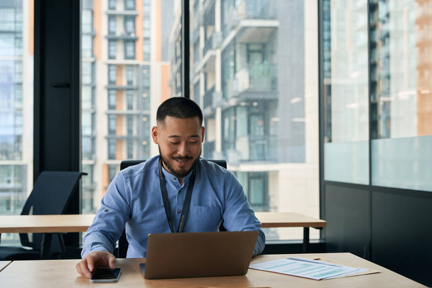 Усміхнений молодий бізнесмен сидить за офісним столом і дивиться на екран свого ноутбука
 - Фото, зображення
