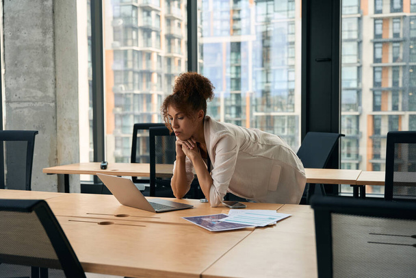 Serious focused businesswoman leaning on office desk and looking at laptop screen - 写真・画像