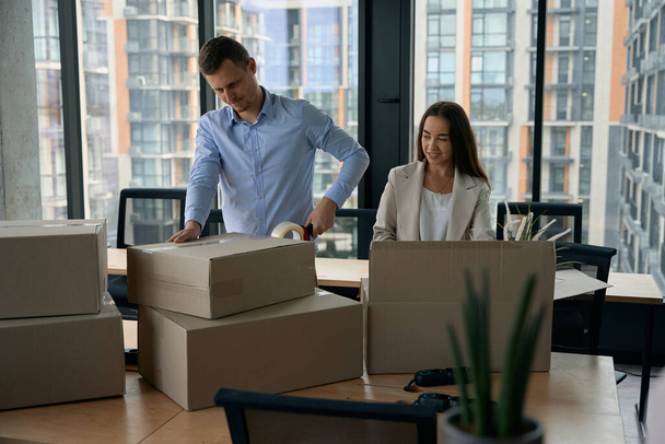 Smiling company employee watching her colleague sealing cardboard box with Scotch tape - Photo, image