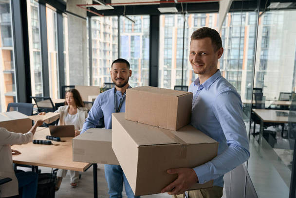Radostní firemní zaměstnanci držící zapečetěné kartonové krabice, zatímco jejich spolupracovníci připravují přepravní doklady - Fotografie, Obrázek